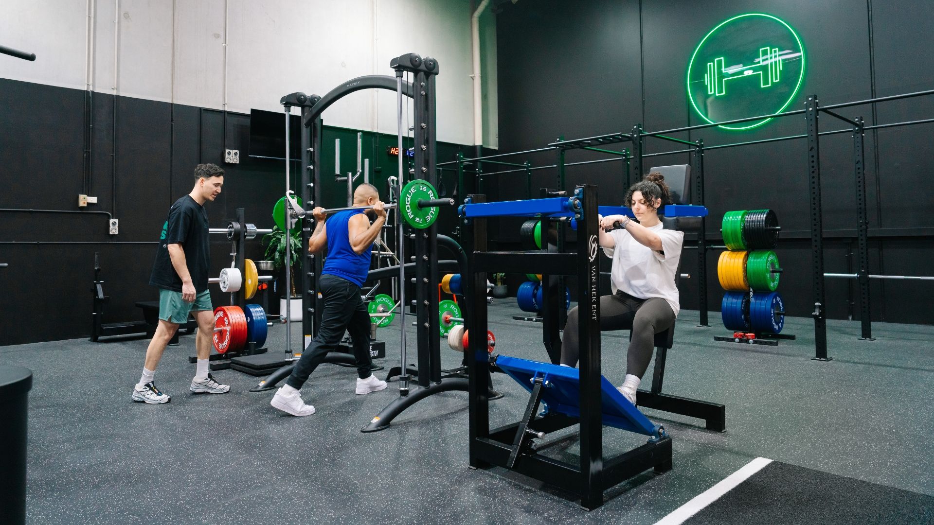 A personal trainer guides a client through a seated dumbbell shoulder press in a modern gym. The client lifts two dumbbells at shoulder height while the trainer stands behind with arms raised, demonstrating proper movement and encouraging correct form.

