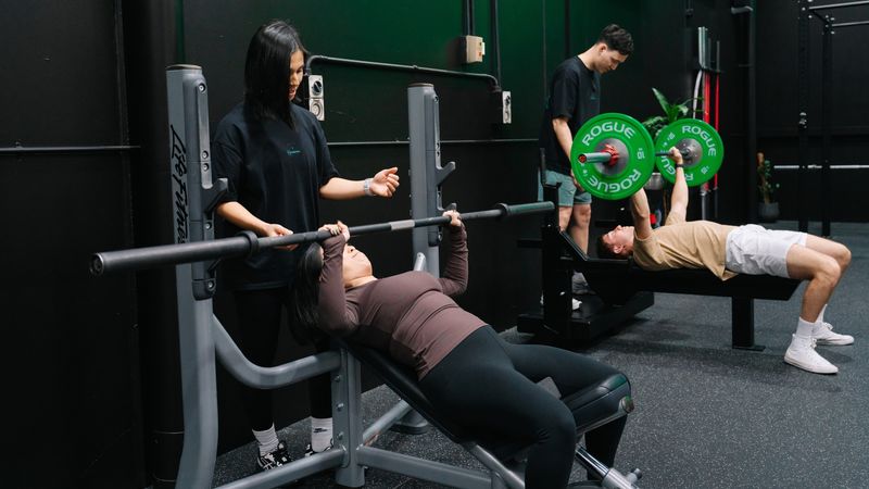 A gym member performs a heavy safety bar squat inside a power rack while a coach watches closely to provide guidance and ensure proper technique. Bright green weight plates and a neon gym logo are visible in the background.