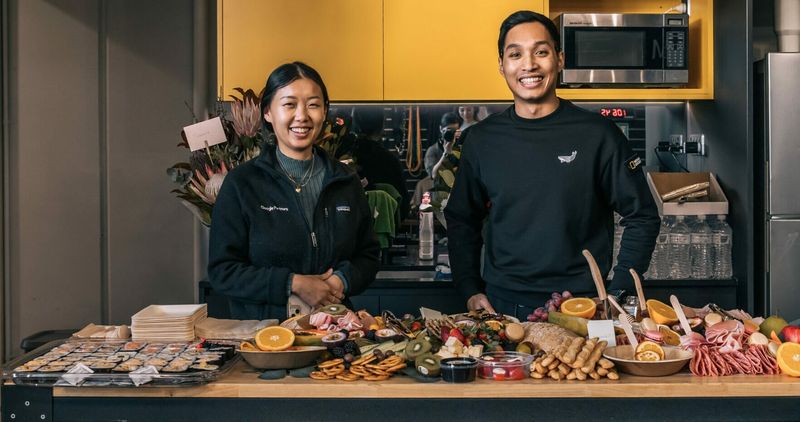 Habitat Gym members posing with a grazing table with fresh fruit, meats and snacks inside the kitchen