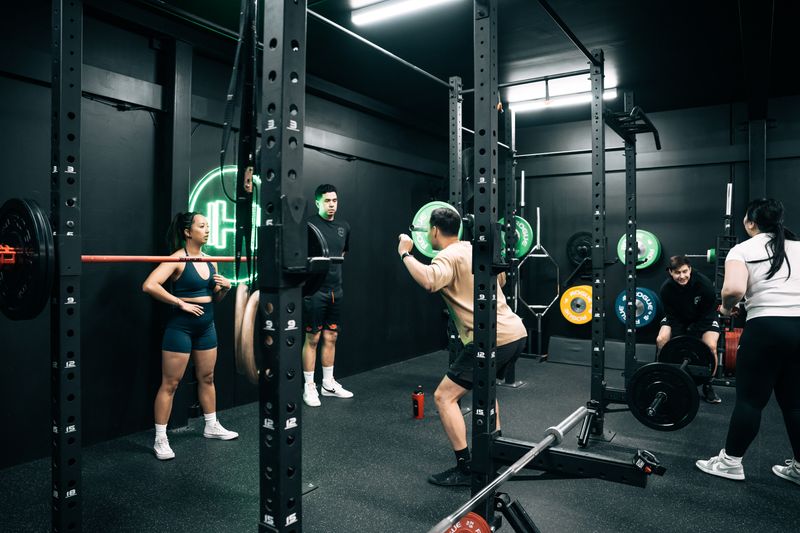A group of people lifting weights at Habitat Gym. One man is performing a barbell back squat inside a squat rack, while others observe or train nearby. The gym has a black interior with neon lighting and coloured Rogue weight plates. A female coach stands to the left, watching attentively.