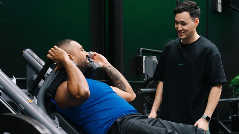 A personal trainer supervises a client performing a strength training exercise on a seated machine at the gym. The client is focused on the movement while the trainer provides guidance and support, highlighting the importance of proper form and personalized coaching.