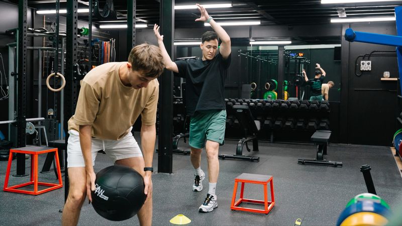 Two men training at Habitat Gym, with one performing a medicine ball lift while a trainer demonstrates proper movement and technique in the background. The gym features strength training equipment, squat racks, and functional fitness tools.