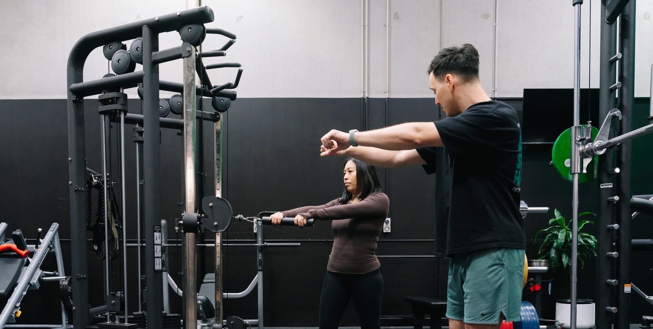 Coach demonstrating the movement while a member practises front raises at the functional trainer during a session at Habitat Gym in Prestons