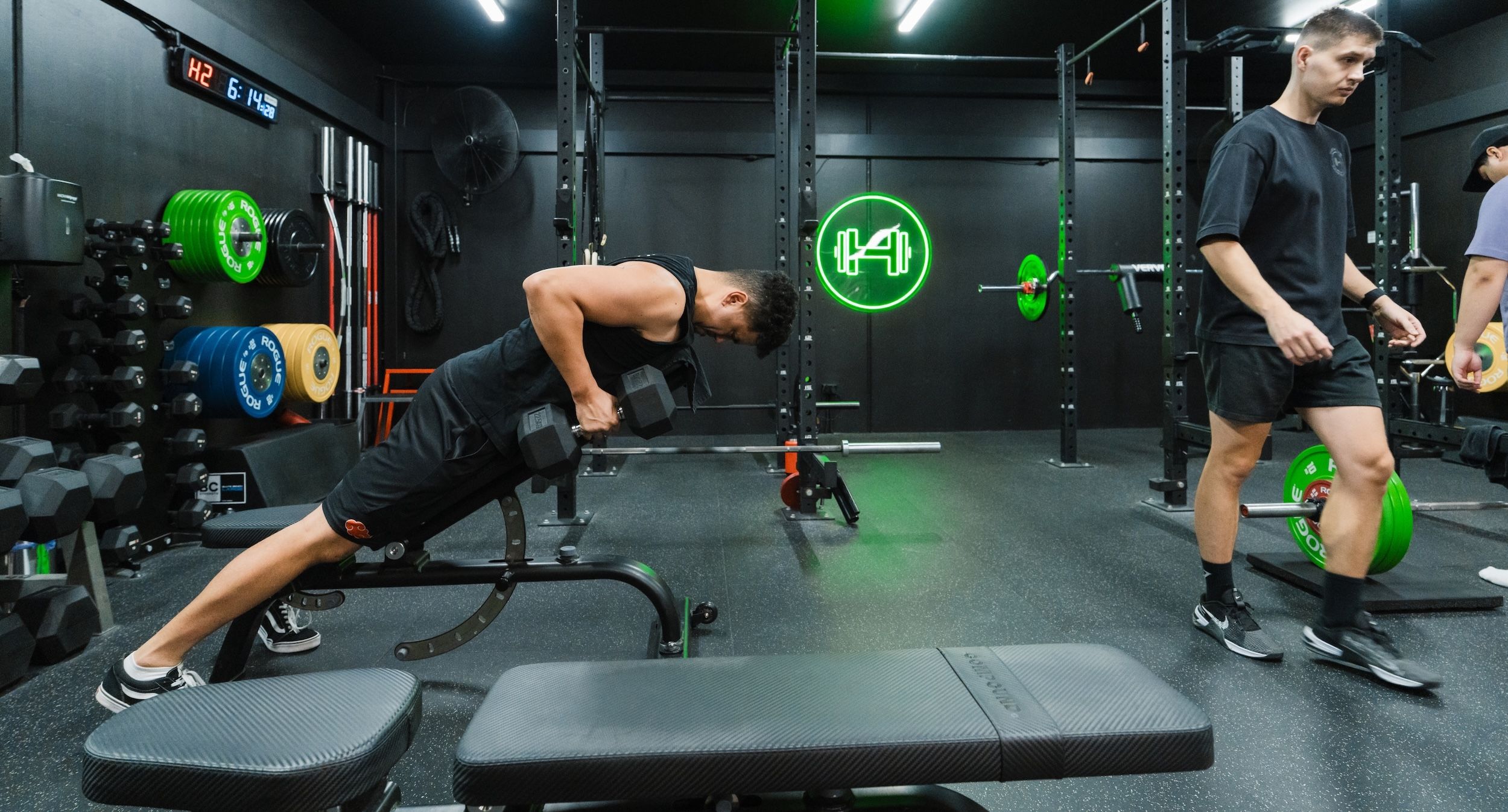 Man in a black tank top performing chest-supported dumbbell rows on an incline bench, while another member walks past in the background. The gym’s green neon dumbbell logo glows on the back wall, with racks of dumbbells and coloured bumper plates lining the space.