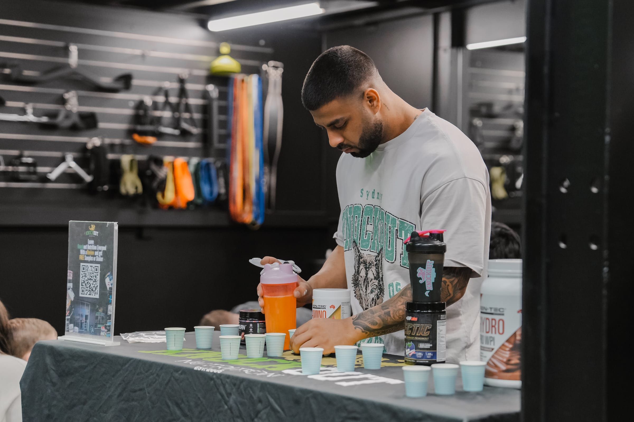 Partner brand rep pouring supplement samples into tasting cups at a Habitat Gym community event, resistance bands displayed on the wall behind