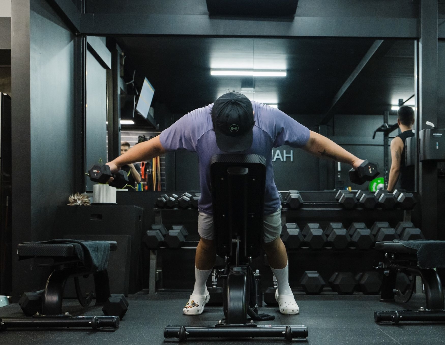 Man in a purple shirt and cap seated on a bench performing bent-over rear delt flys with dumbbells, viewed from behind. A coach in a black tank top stands to the side, smiling as he watches the lift. Dumbbell rack and Rogue plates fill the background.