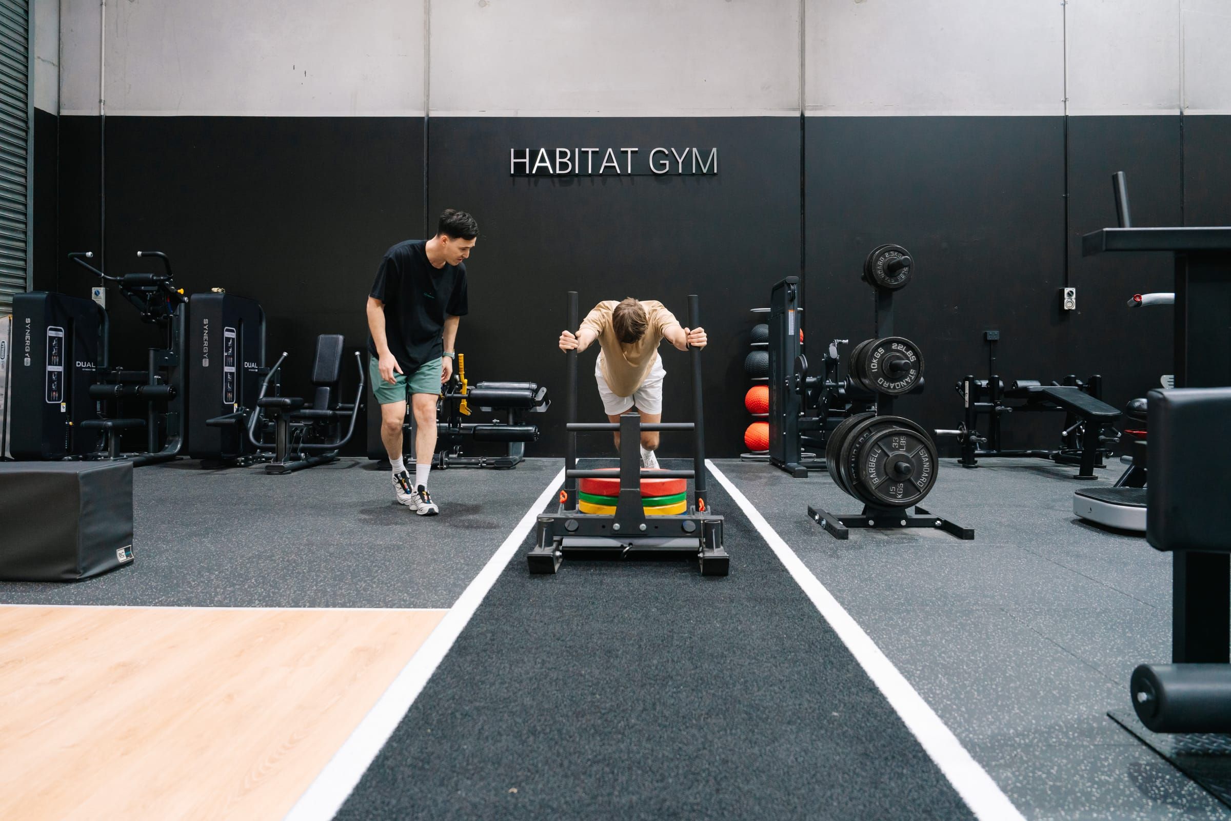 Member driving a loaded sled down the track as a coach runs alongside during a strength and conditioning session at Habitat Gym in Prestons