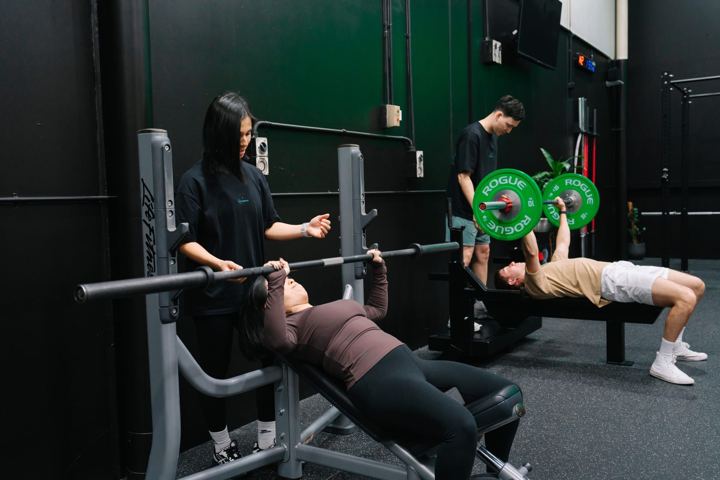 Coach guiding a member through an incline bench press while another member benches with Rogue plates in the background at Habitat Gym in Prestons