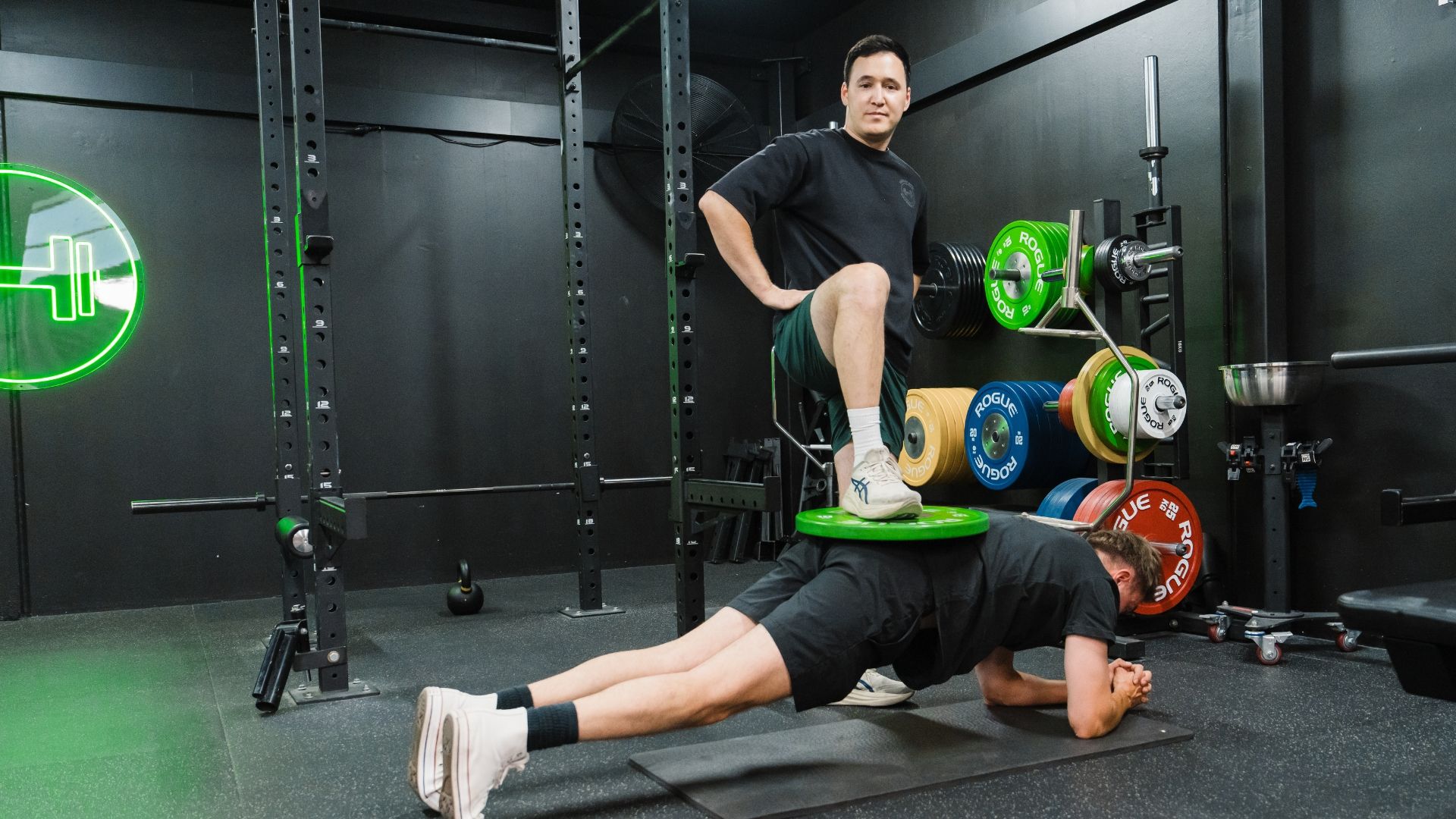 Personal trainer supervising weighted plank exercise inside strength training gym with squat rack and bumper plates