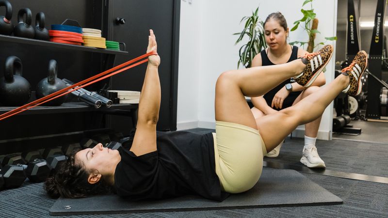 Coach guiding client performing resistance band core exercise on gym floor with kettlebells and equipment in the background
