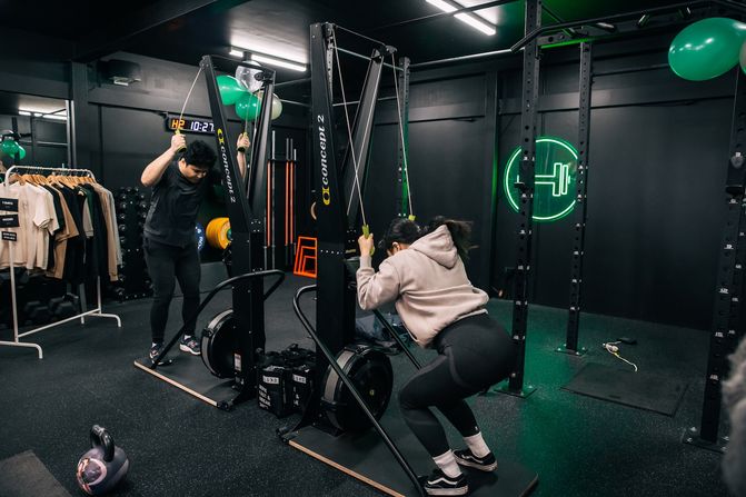 Two members training side by side on Concept 2 SkiErg machines, one standing tall mid-pull and the other dropping into a squat at the bottom of the stroke. A green neon dumbbell sign glows on the back wall.