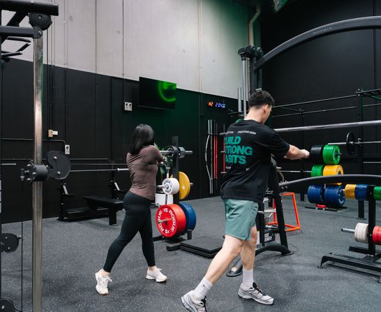 Two members moving through barbell work in Habitat Gym’s strength zone, coloured bumper plates racked behind them