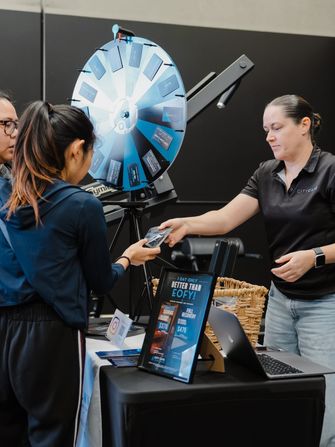 Stallholder helping two visitors with a card reader beside a spin-to-win prize wheel at a Habitat Gym partner event