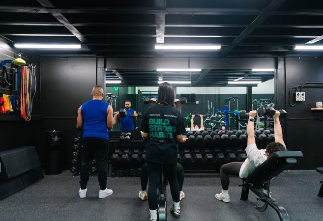 Coach and members mid-session on the dumbbell floor, one pressing dumbbells from a bench in front of the mirror