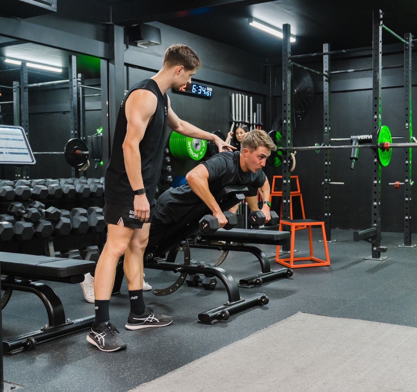 Coach in a black tank top guiding a member through single-arm dumbbell rows on an incline bench, with another member doing floor work near the rack in the background. A green neon dumbbell sign glows on the wall.