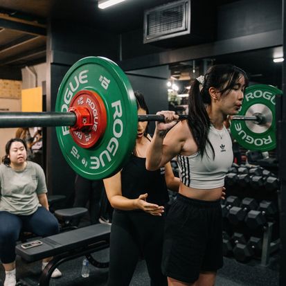 Woman performing a barbell squat during a strength training workshop at a gym, with a trainer providing guidance and another participant observing. The session focuses on proper lifting technique, strength and fitness.
