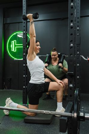 Woman in a white tank top kneeling on a pad in a power rack, pressing a dumbbell overhead with one arm while another woman in a green shirt squats behind her. A green neon dumbbell sign (Habitat Gym's logo) glows on the wall in the background.