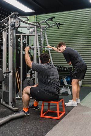 Man seated on a red plyo box performing a single-arm cable pulldown on a Life Fitness machine, while a trainer in a dark grey shirt stands beside him guiding his technique.